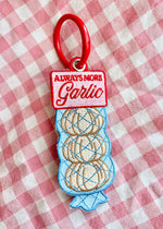 an embroidered keychain with a chunky red key ring. The design features a pack of garlic bulbs in mesh. The label reads "always more GARLIC". this would be a perfect gift and accessory or trinket for anyone who loves to cook!