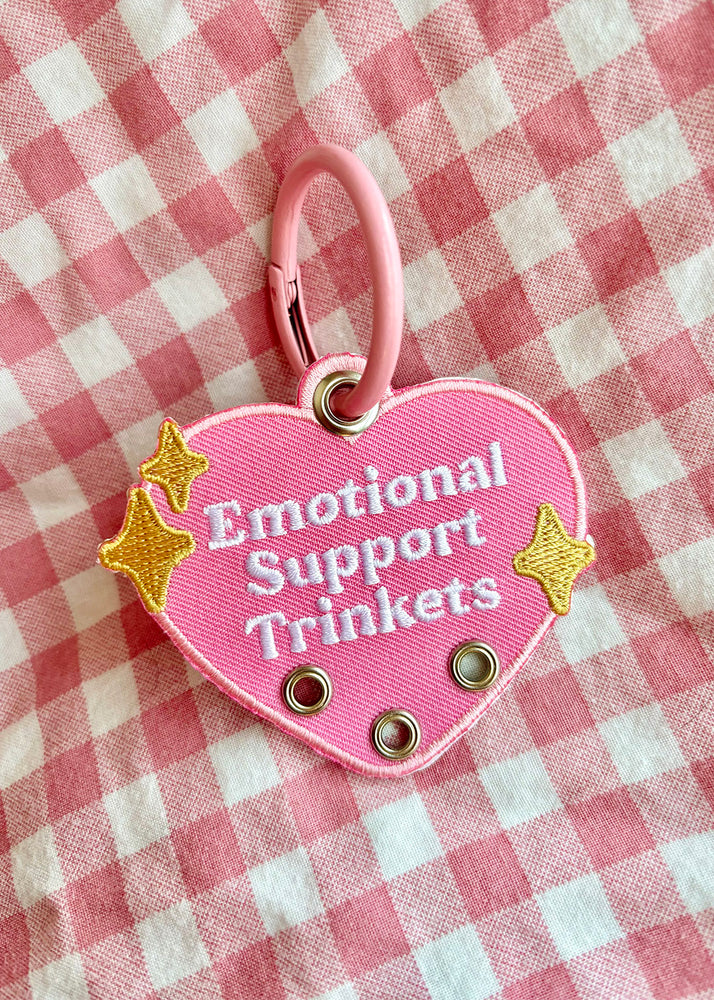 an embroidered patch keychain with a chunky colorful metal key ring. The funny and cute heart shaped design features 3 eyelet holes that allow you to attach more keychains or trinkets. This cute pink purse charm is bright and colorful and features gold sparkles surrounding the text " Emotional Support Trinkets". 