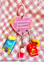 an embroidered patch keychain with a chunky colorful metal key ring. The funny and cute heart shaped design features 3 eyelet holes that allow you to attach more keychains or trinkets. This cute pink purse charm is bright and colorful and features gold sparkles surrounding the text " Emotional Support Trinkets". This image shows the keychain with lots of gachapon trinkets and charms attached to it.
