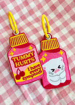 an embroidered patch keychain with a chunky colorful metal key ring. The funny and cute design is shaped like a pepto bismol bottle and reads TUMMY HURTS I have every symptom. The flavor is cherry and the bottle is very pink and bright and fun. This would be a perfect little accessory for someone who has chronic tummy or digestion issues. On the bag, there is a cute crying white cartoon cat holding its tummy.